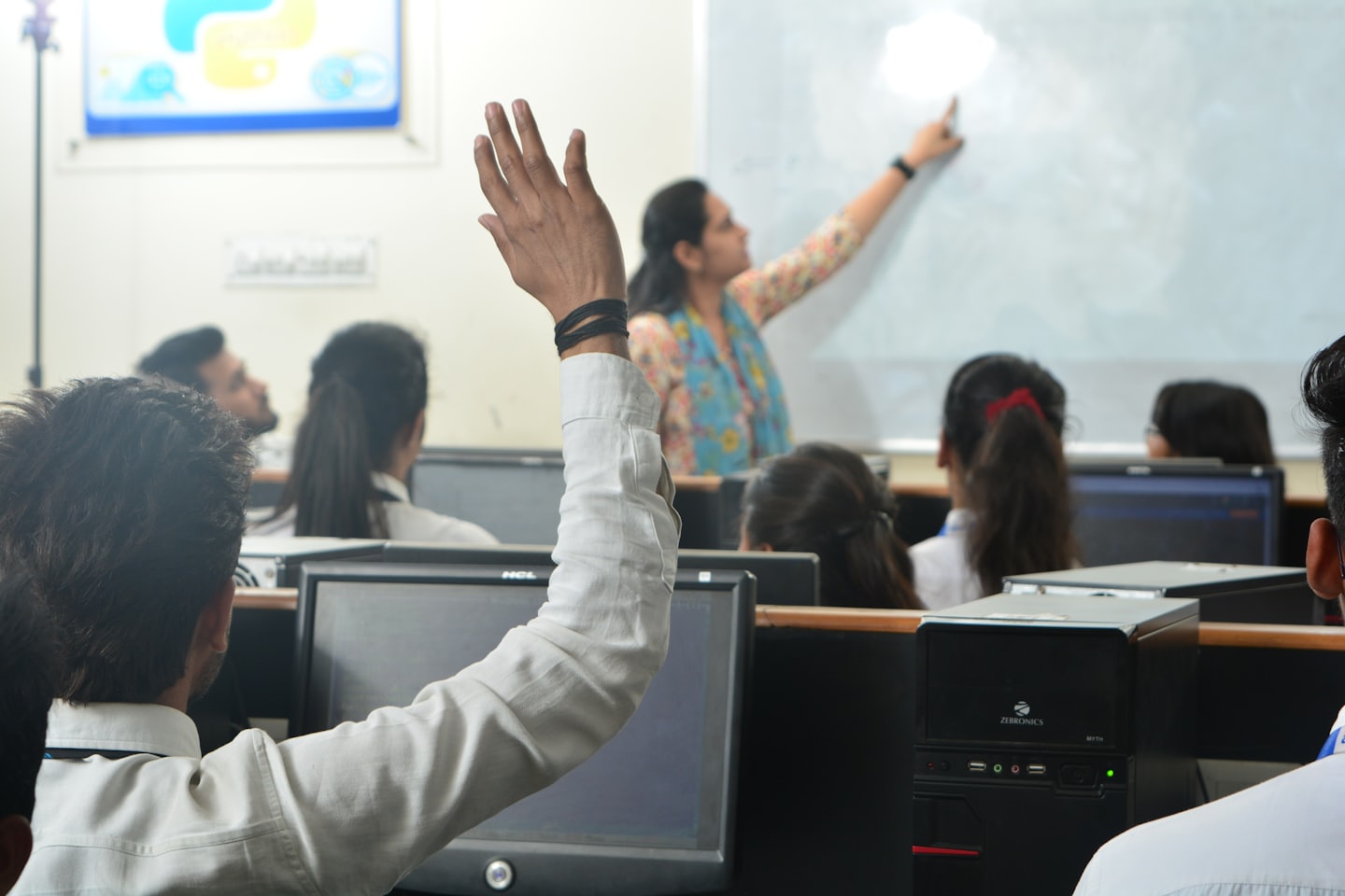 Teacher points at whiteboard in a busy computer lab classroom.